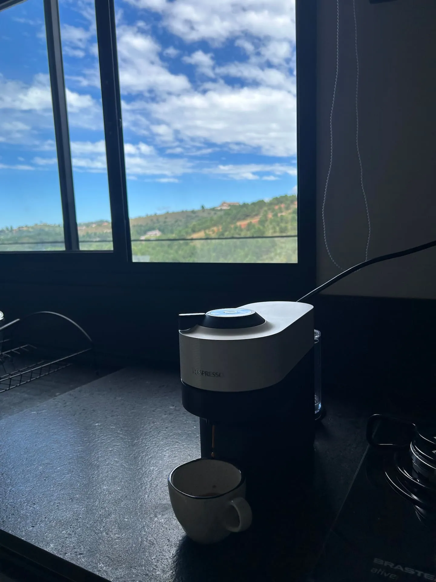 Cafeteira e xícara na bancada, com céu azul na janela