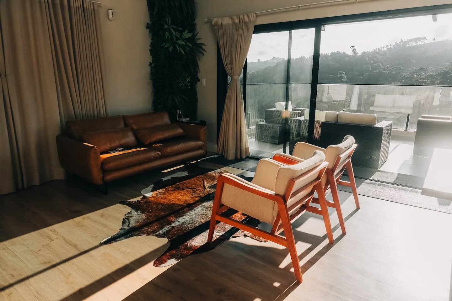 Sala com sofá de couro, poltronas e luz do sol entrando pelas portas de vidro