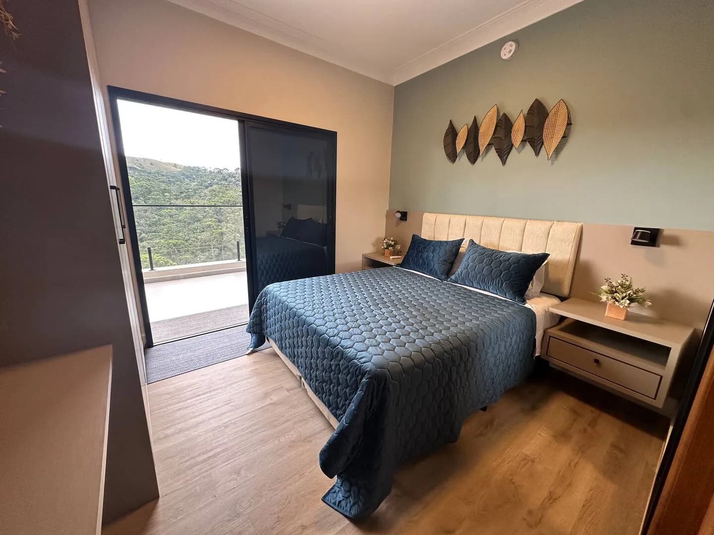 Suíte com cama queen, colcha azul e porta de vidro para a varanda